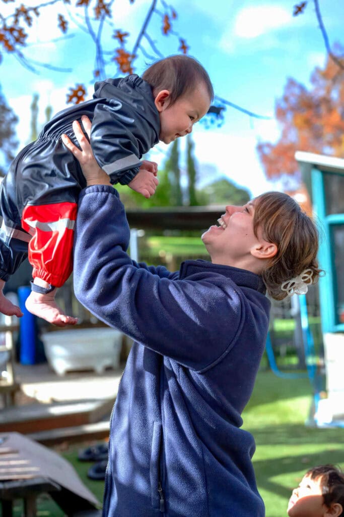 The kind teacher picks up the one-year-old child in a childcare playground