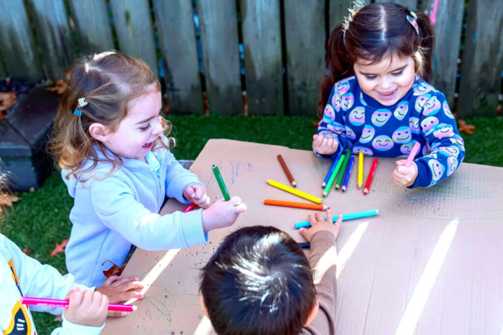 Childcare children drawing big picture on cardboard sharing pencils together